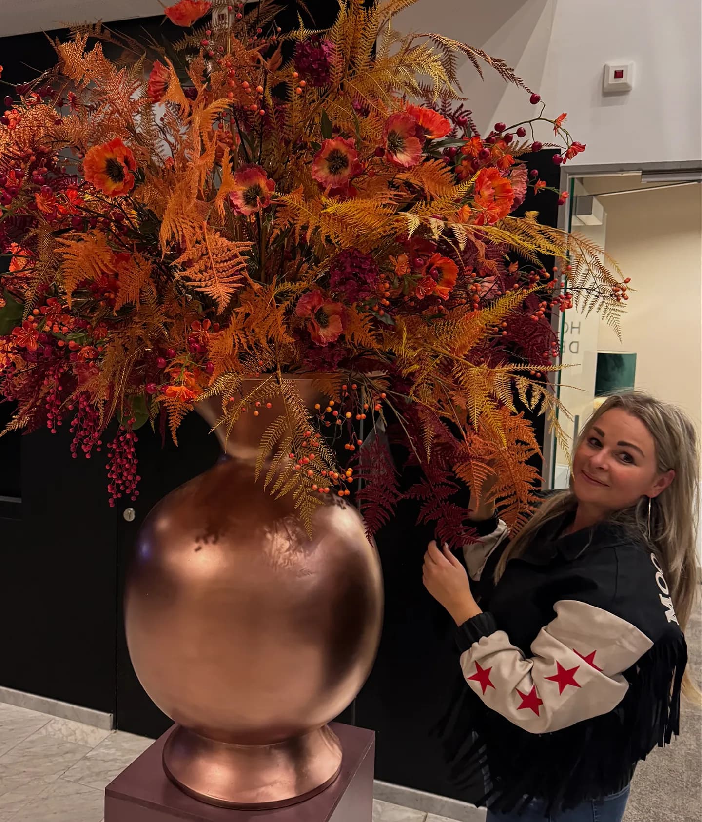 Enorm herfstboeket in een ronde koperkleurige vaas, met een lachende vrouw ernaast